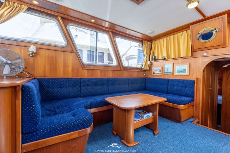 Een interieur van een boot met houten wanden, een blauwe bank, een houten tafel en ramen met gele gordijnen.