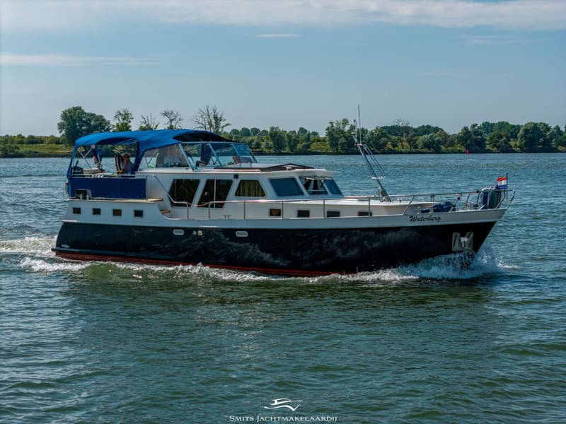 Een donkerblauwe motorboot met een witte opbouw en blauwe bimini vaart op een rivier met bomen op de achtergrond.