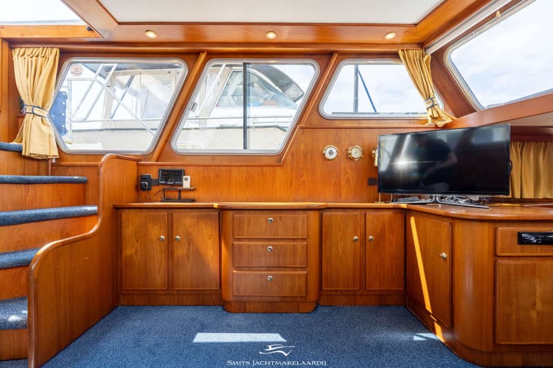Een interieur van een boot met houten wanden, een blauw tapijt, ramen met gele gordijnen, een trap en een televisie.