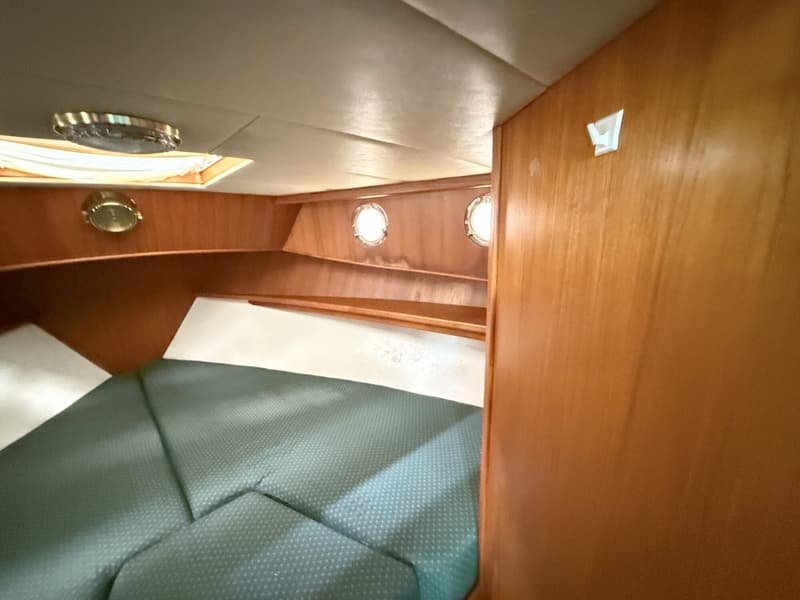 De foto toont een kajuit met een groen gestoffeerd bed, houten wanden, twee patrijspoorten en een plafondlamp.
