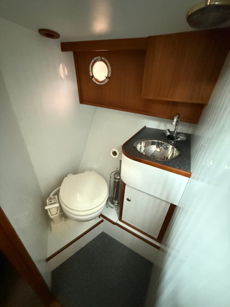 Een bootbadkamer met een wit toilet, een ronde wasbak met kraan, een patrijspoort en een douchekop aan het plafond.