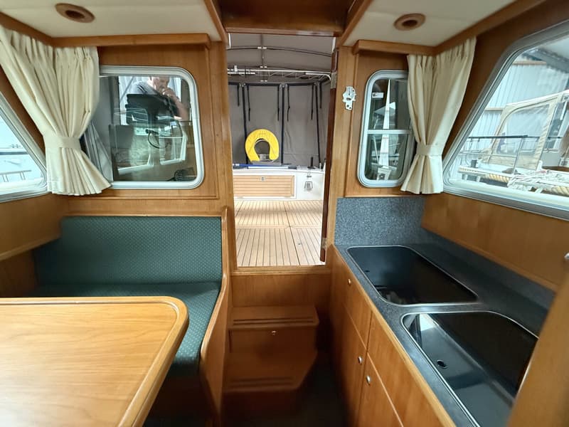 De foto toont het interieur van een boot met een houten tafel, groene bank, ramen met gordijnen, een keukenblok en een…