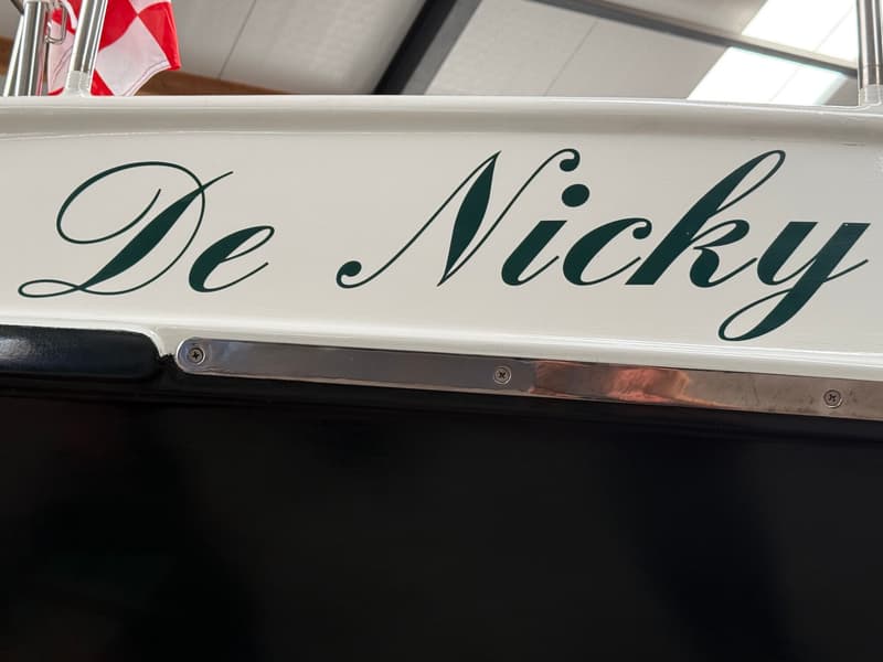 De naam "De Nicky" staat in donkergroene sierletters op een witte romp, met een rood-wit geblokte vlag erboven.