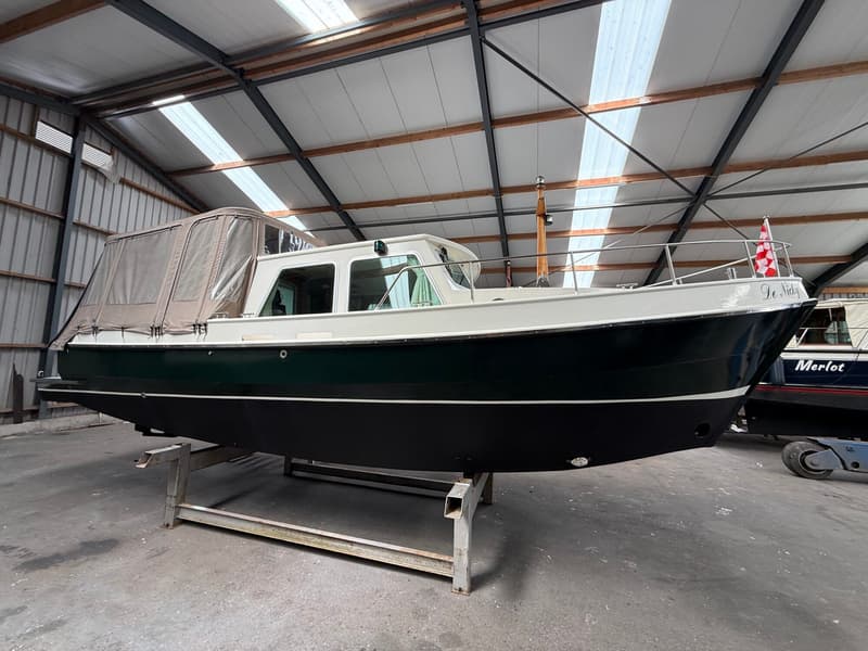 Een donkergroene boot met een witte opbouw en een beige afdekzeil staat op een metalen bok in een loods.