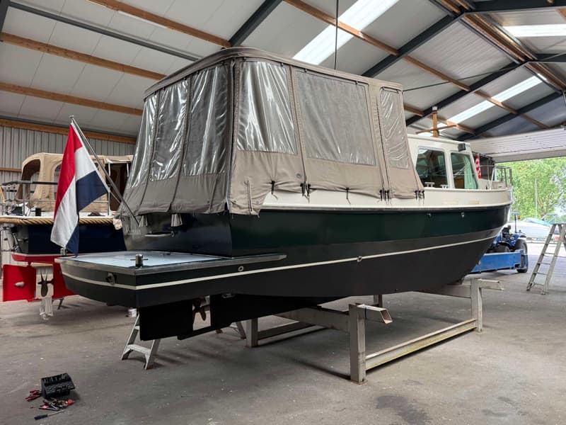 Een donkergroene boot met een beige afdekzeil staat op bokken in een loods. — Barkas Barkas 8.30 OK