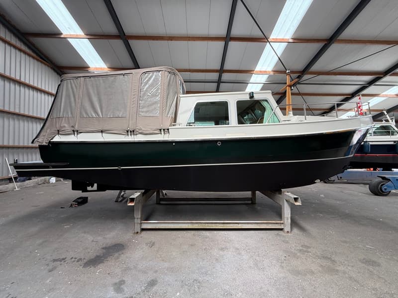 Een donkergroene boot met een witte opbouw en een beige cabriokap staat op een metalen bok in een loods.