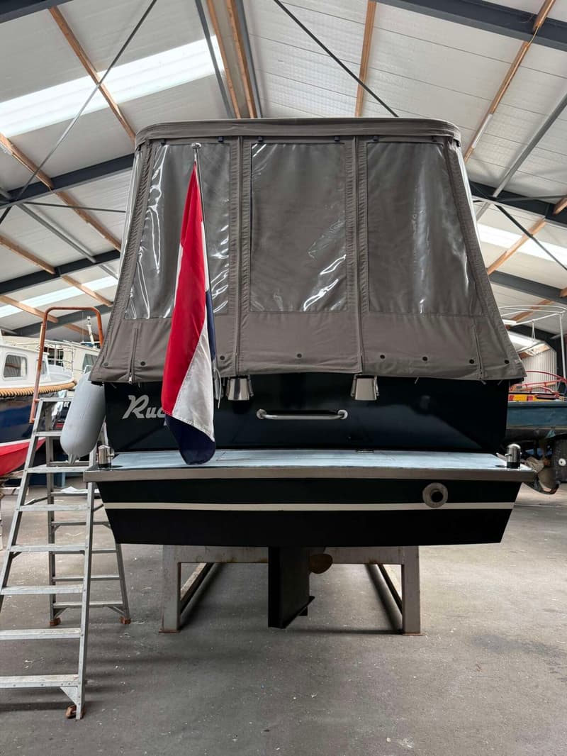 De achterkant van een donkerblauwe boot met een grijze cabriokap en een Nederlandse vlag, staand op een trailer in een loods.