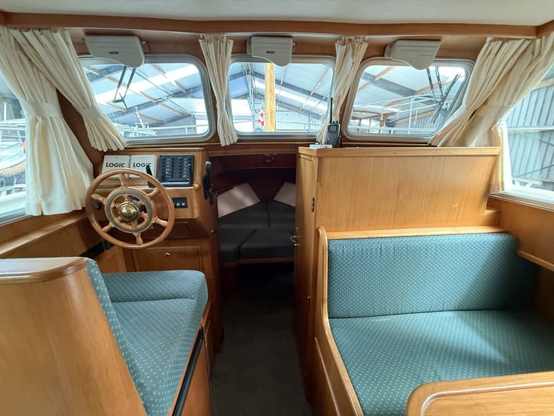 De foto toont het interieur van een boot met een houten stuurwiel, groene gestoffeerde banken en een doorkijk naar een…