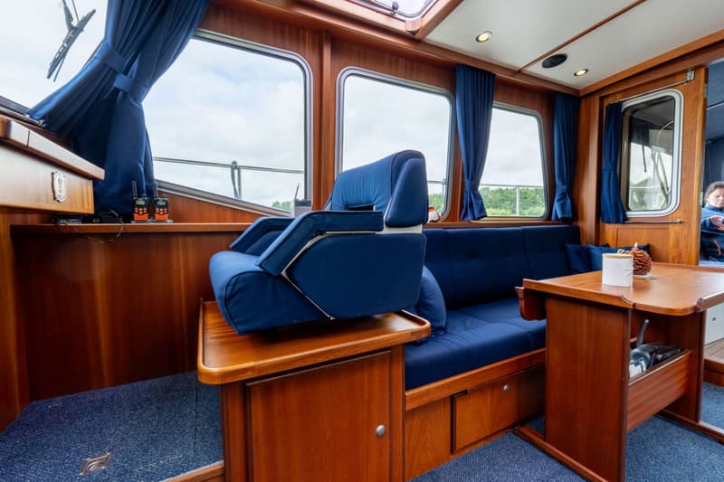 Het interieur van een boot met houten wanden, blauwe bekleding, een tafel en ramen met uitzicht naar buiten.