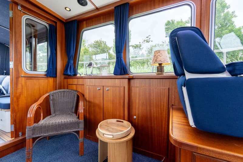 De foto toont een interieur van een boot met houten wanden, ramen met blauwe gordijnen, een rieten stoel en een blauwe stoel.