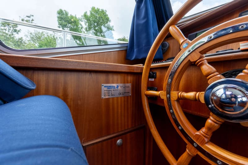 Een houten stuurwiel met chromen details, een blauwe stoffen zitting en een typeplaatje van Brandsma Jachten.