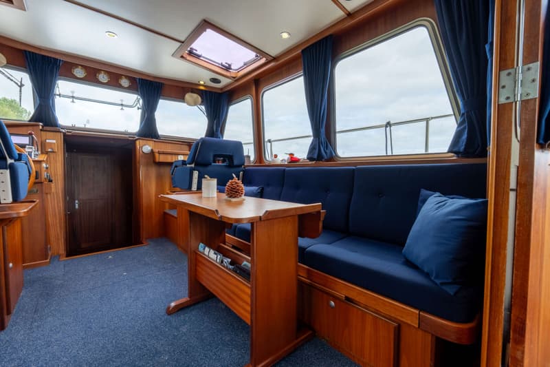 De foto toont het interieur van een boot met een blauw tapijt, houten lambrisering, blauwe banken, een houten tafel en…