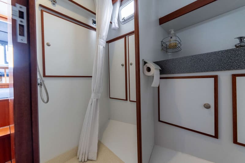 Een witte doucheruimte met houten accenten, een douchekop, een raam, een toiletrolhouder en opbergkasten.
