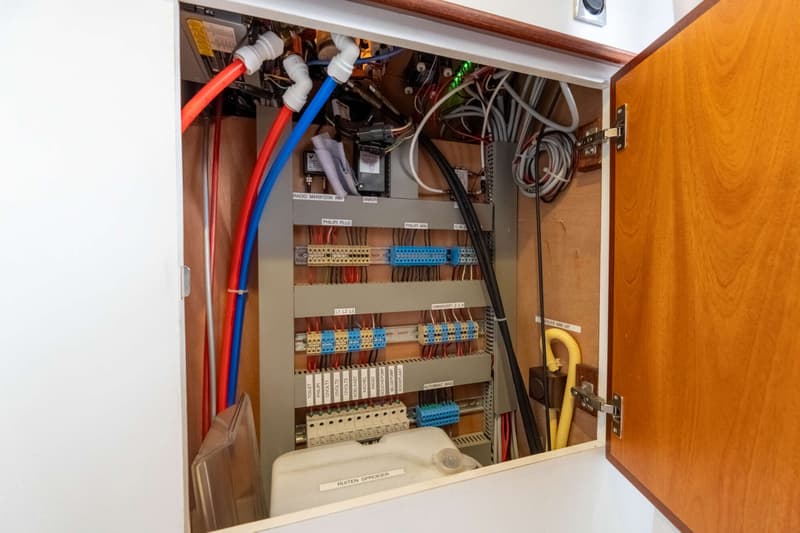 Een open kast met een elektrisch paneel, bedrading, rode en blauwe leidingen, en een wit reservoir.