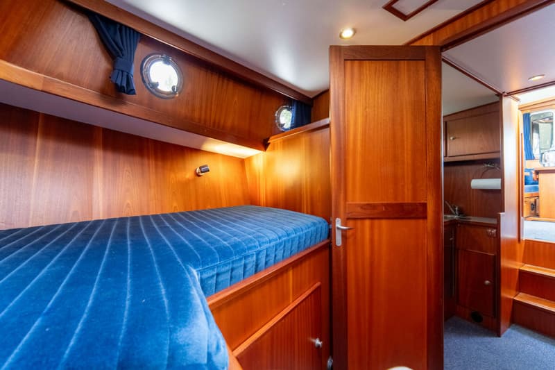 De foto toont een kajuit met een bed met blauw gestoffeerd matras, houten wanden en twee patrijspoorten.