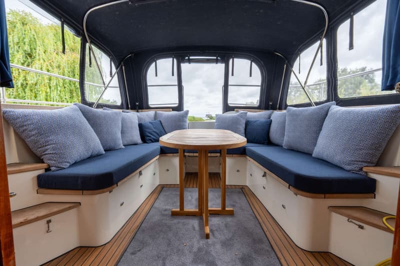De foto toont een interieur van een boot met een U-vormige zithoek, een houten tafel en een donkerblauwe kap.