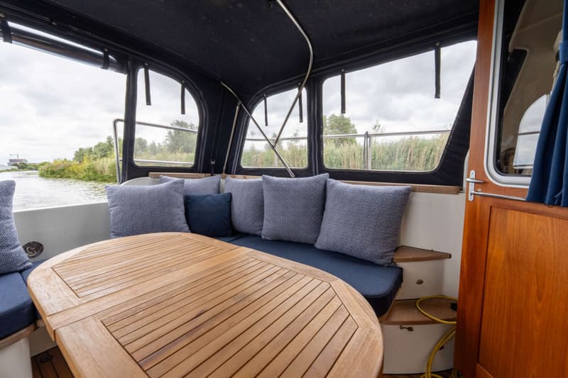 Een houten tafel met latten staat voor een bank met blauwe kussens en sierkussens, met uitzicht op water en riet.