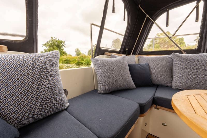 De foto toont een bootinterieur met blauwe kussens en een houten tafel, met uitzicht op groen langs het water.