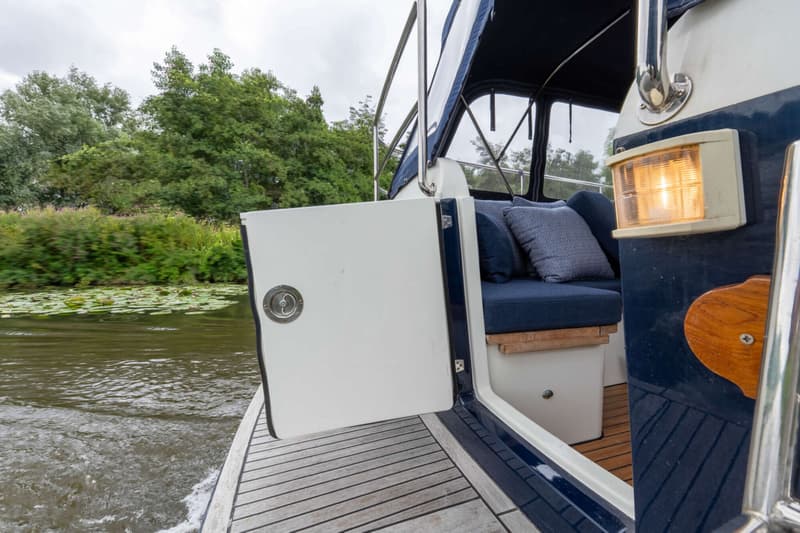 Een boot met een open deur en een verlicht navigatielicht vaart langs een oever met waterlelies en bomen.
