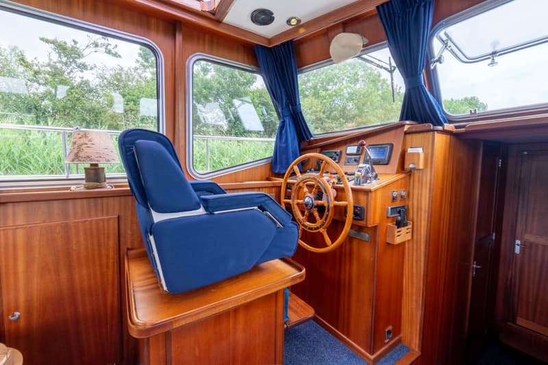 Een interieur van een boot met een houten stuurwiel, een blauwe stoel en ramen met uitzicht op groen.
