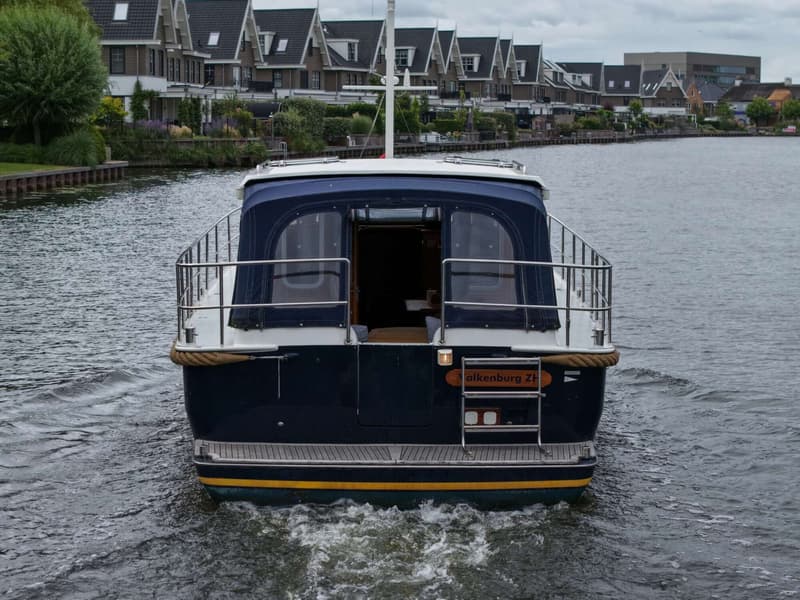 Een donkerblauwe boot met een witte opbouw en een blauwe kap vaart over het water, met huizen op de achtergrond.