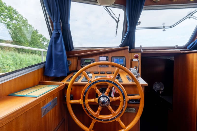 De foto toont het interieur van een stuurhut met een houten stuurwiel, diverse navigatie-instrumenten en ramen met blauwe…