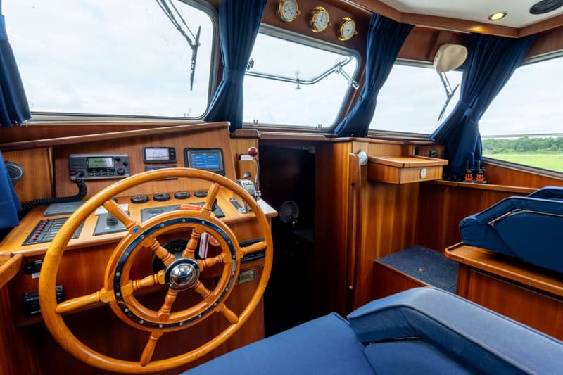 De foto toont het interieur van een stuurhut met een houten stuurwiel, navigatieapparatuur en blauwe kussens.