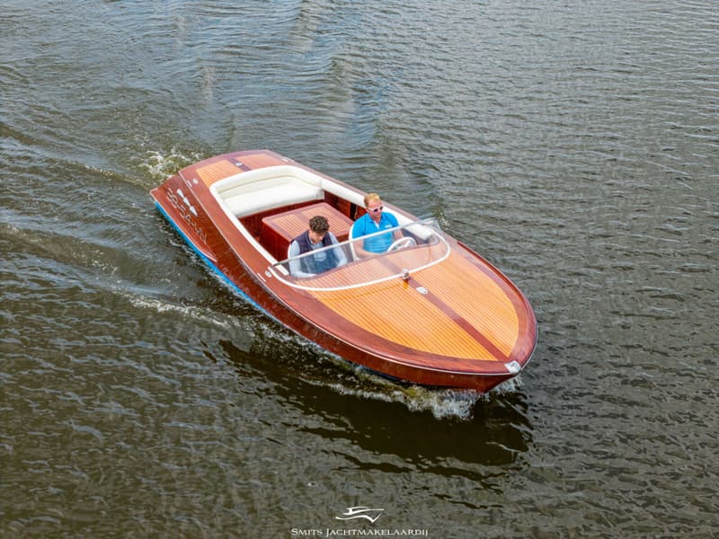 Een houten motorboot met twee mannen aan boord vaart over water, met registratienummer 38-54-YN op de zijkant.