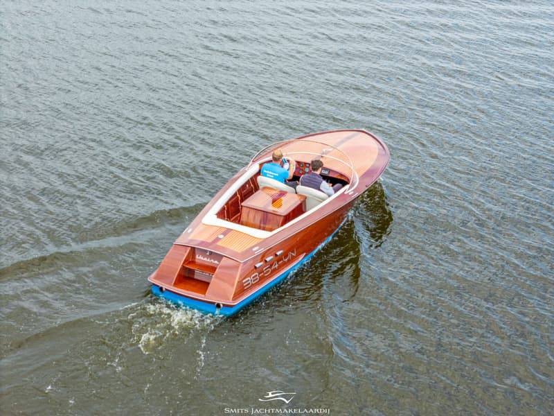 Een houten motorboot met twee personen vaart over het water, met registratienummer 38-54-YN op de zijkant.