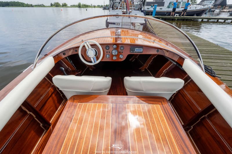 De foto toont het interieur van een houten boot met een wit stuur, witte stoelen en een dashboard met meters.