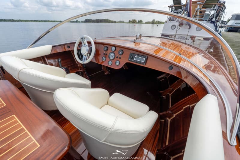 Een houten boot met witte stoelen en een wit stuur, liggend aan een steiger met water en bomen op de achtergrond.