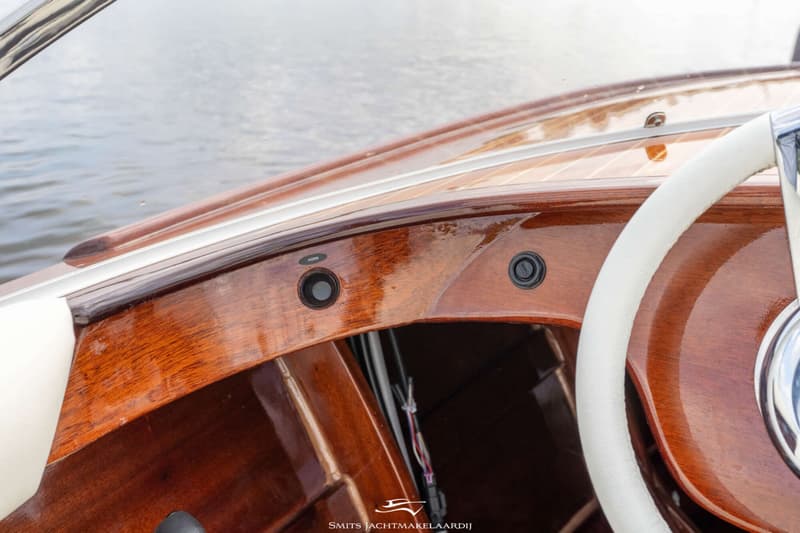 Een detailfoto van een glimmend houten dashboard met een wit stuurwiel en twee zwarte knoppen, waarvan één met "HORN".