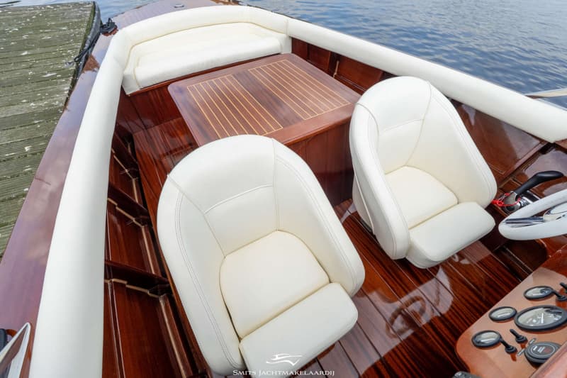Een bovenaanzicht van de cockpit van een houten boot met witte bekleding, een houten tafel en een stuur.