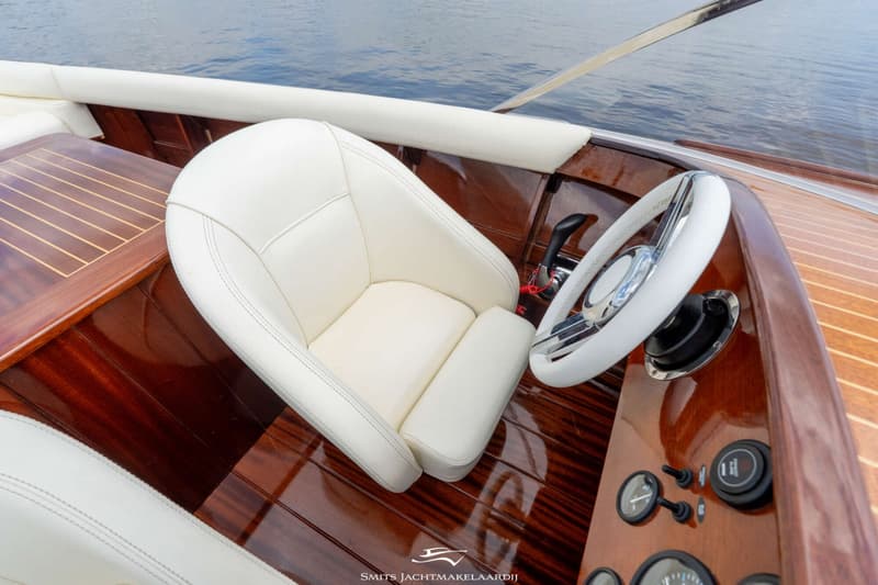De foto toont een interieur van een boot met een wit stuur, een witte stoel, een houten dashboard en een houten vloer.