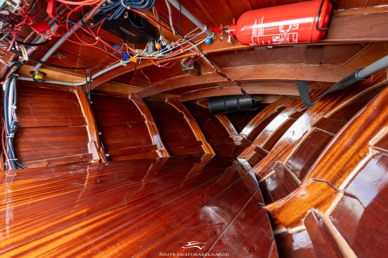 De foto toont het interieur van een boot met glanzend gelakt hout, zichtbare spanten, een brandblusser en wirwar van kabels.