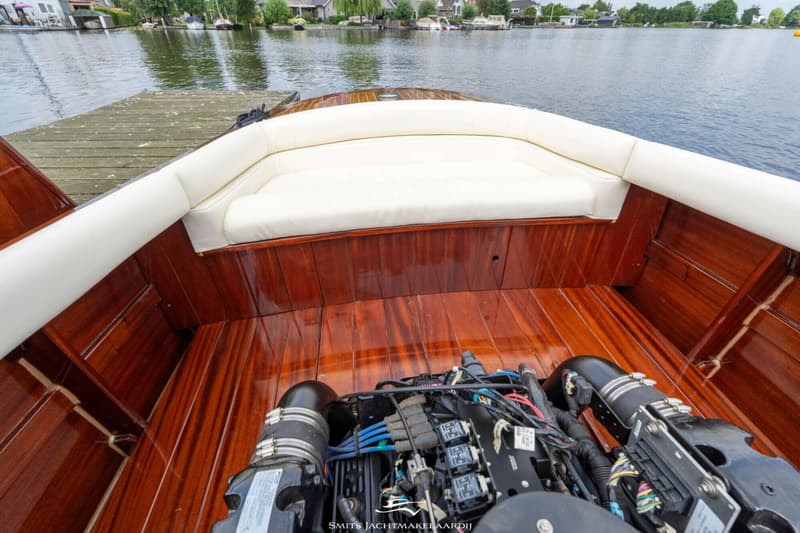 De foto toont de achterkant van een boot met een glanzende houten afwerking, witte kussens en een motor.