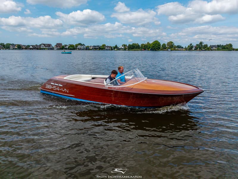 Een houten speedboot met twee mannen aan boord vaart over een meer met huizen aan de oever onder een bewolkte hemel.