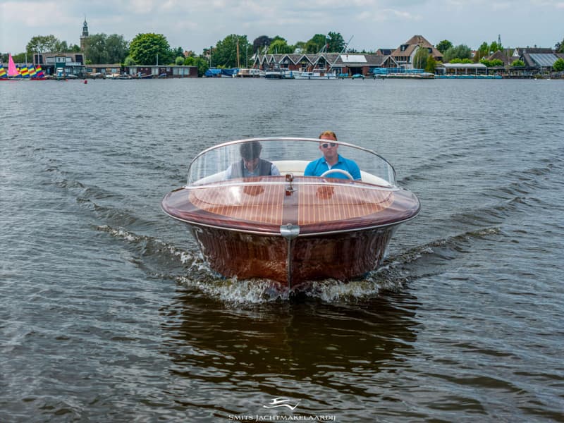 Een houten motorboot met twee mannen aan boord vaart over het water, met een dorp op de achtergrond.