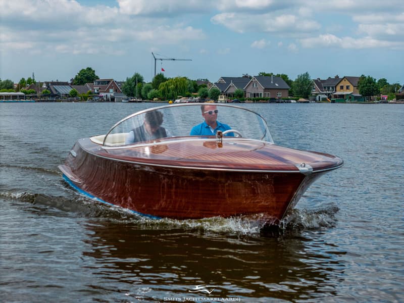 Een houten speedboot met twee mannen aan boord vaart over het water, met huizen en bomen op de achtergrond.