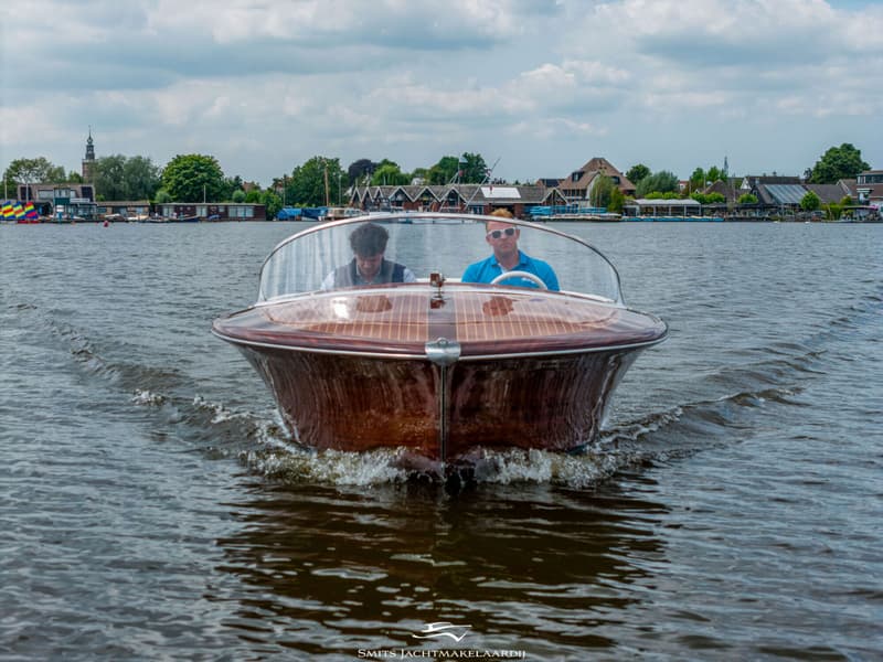 Een houten motorboot met twee mannen aan boord vaart over het water, met een dorp op de achtergrond.