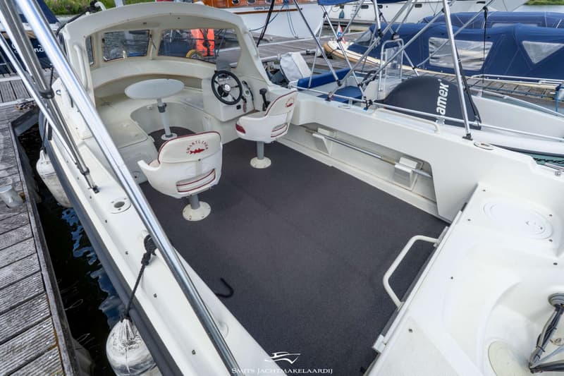 Een witte boot met een donkergrijze vloer, twee witte stoelen, een stuur en een tafel, liggend aan een houten steiger.