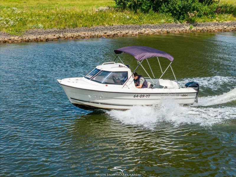 Een witte motorboot met een paarse biminitop vaart over het water, met een persoon aan boord.