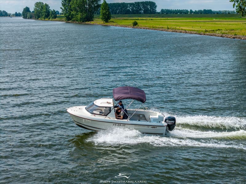 Een witte motorboot met een donkerblauwe biminitop vaart op een rivier, met twee personen aan boord.
