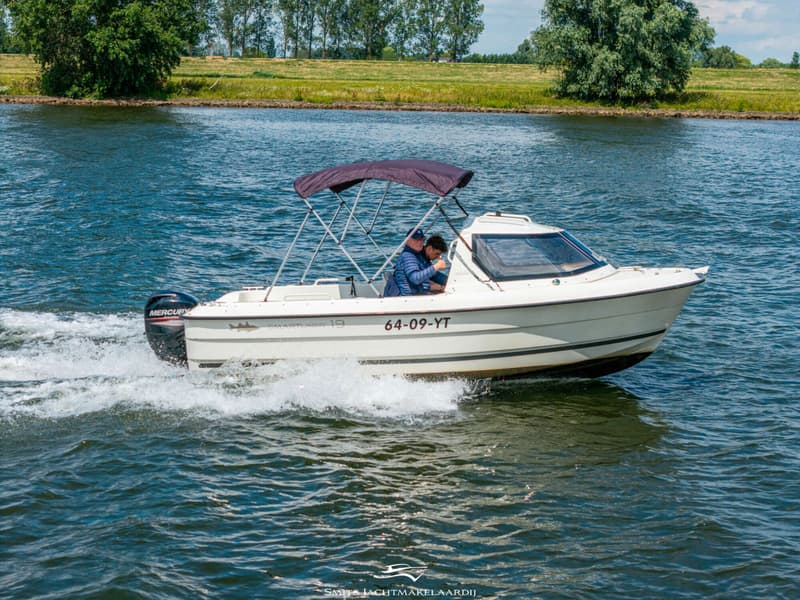 Een witte motorboot met twee personen aan boord vaart over het water, met een groene oever op de achtergrond.