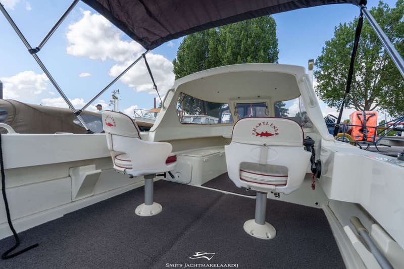 De foto toont het interieur van een boot met twee witte stoelen en een grijs tapijt, met een overkapping en bomen op de…