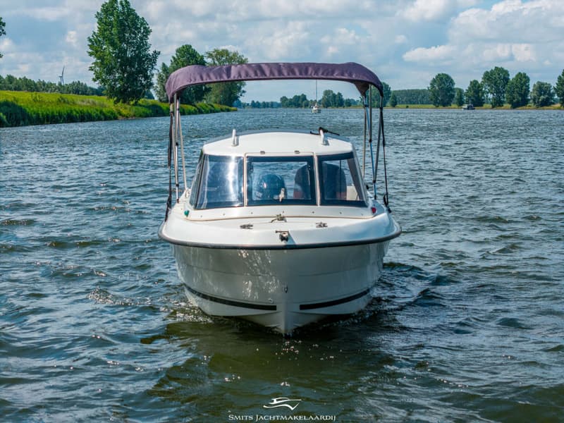 Een witte motorboot met een donkerblauwe biminitop vaart op een waterweg met groene oevers en bomen onder een bewolkte hemel.