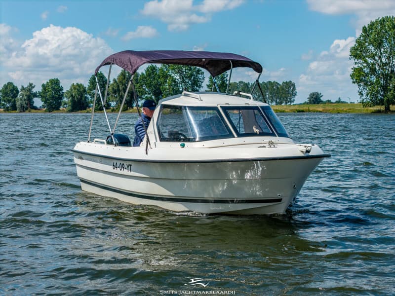 Een witte motorboot met een donkerblauwe biminitop vaart op een waterweg met een man aan het roer.