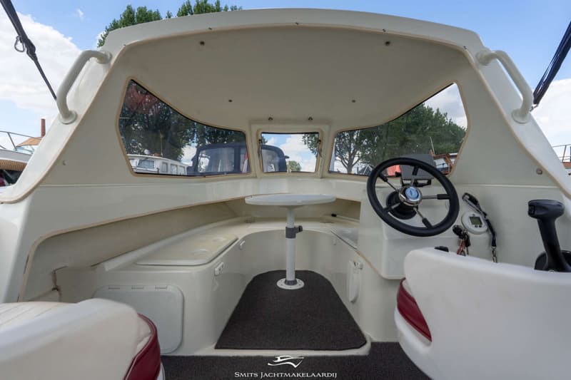 De foto toont het interieur van een boot met een stuurwiel, een tafel en zitplaatsen, gezien vanuit de achterkant.