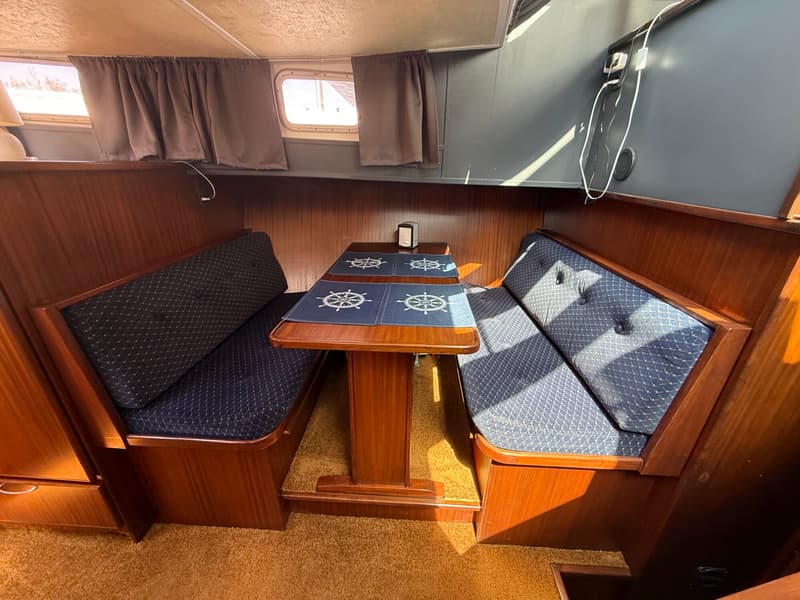 De foto toont een kajuit met een houten tafel en twee banken met blauwe kussens, omringd door houten panelen.