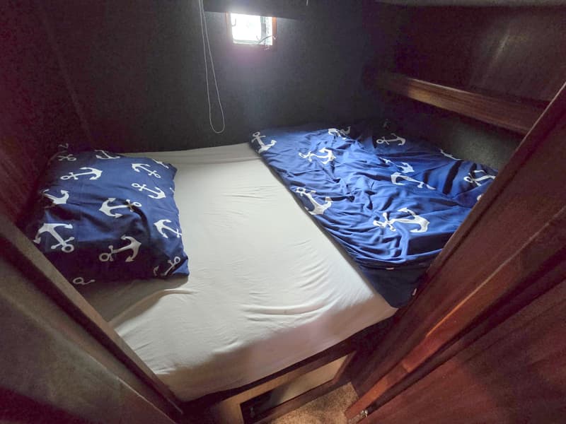Een kajuit met een bed met wit hoeslaken, een blauw dekbed en kussen met ankerpatroon, en een klein raam.
